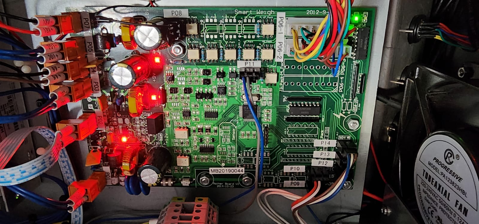A close-up of an open electronic device showing a green circuit board with various components including capacitors, resistors, connectors, and wires. Several red LEDs are illuminated. On the right, a fan is partially visible, suggesting a cooling mechanism.